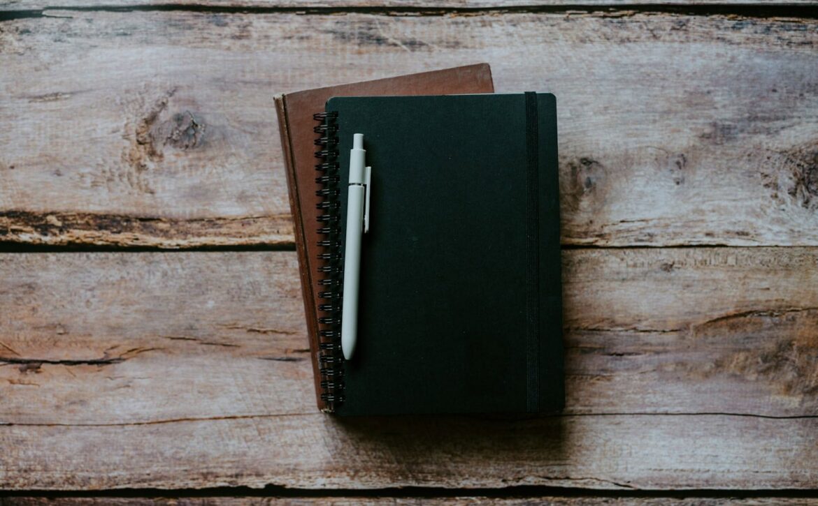 Two closed notebooks and a light-coloured pen on a wooden surface.