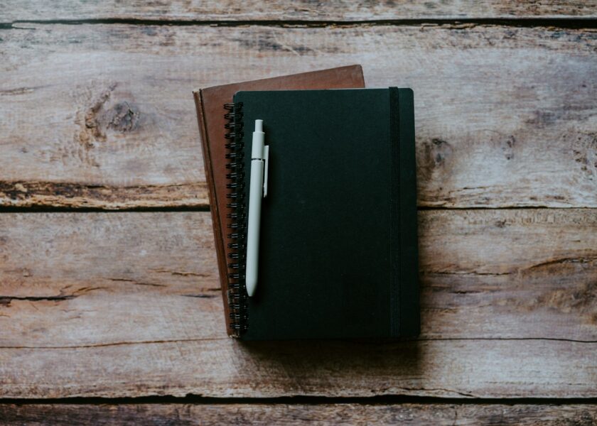 Two closed notebooks and a light-coloured pen on a wooden surface.