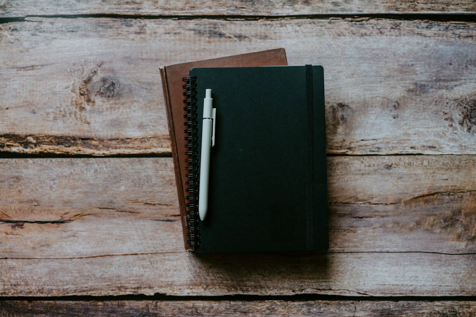 Two closed notebooks and a light-coloured pen on a wooden surface.