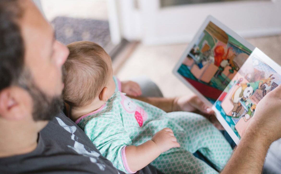 An adult holds an open picture book while a baby rests in their arms.
