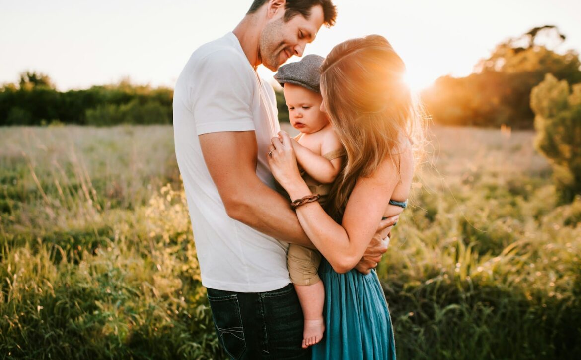 Two adults holding a young child together in a sunlit grassy field.