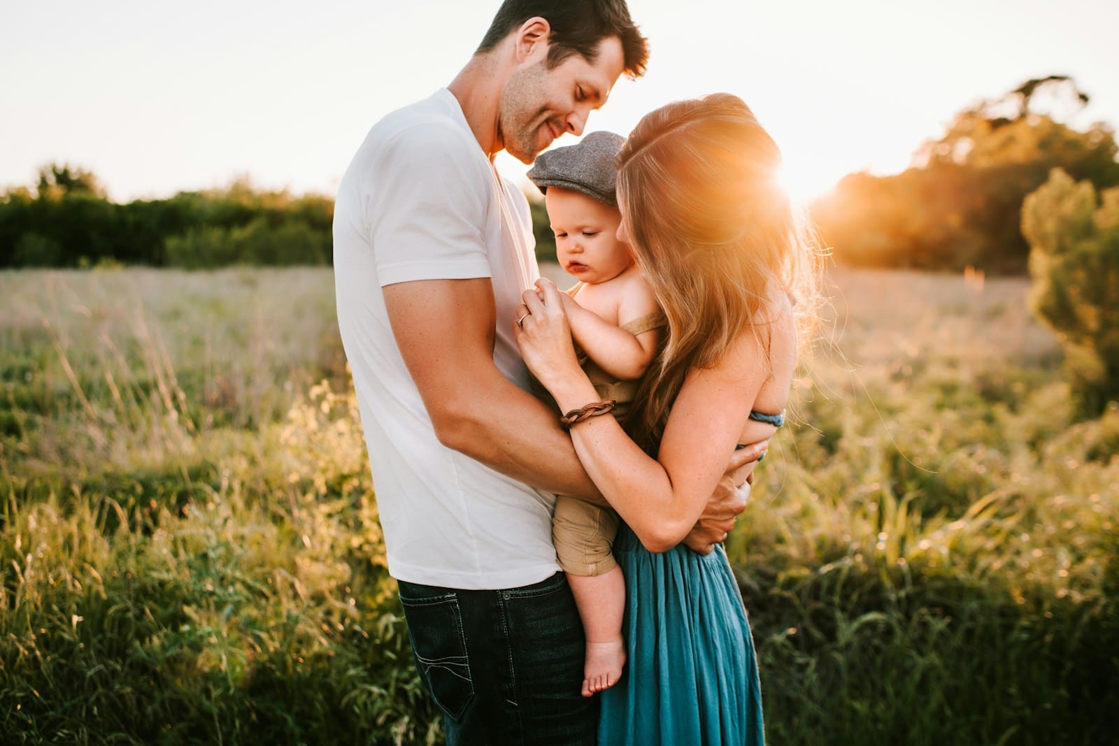 Two adults holding a young child together in a sunlit grassy field.