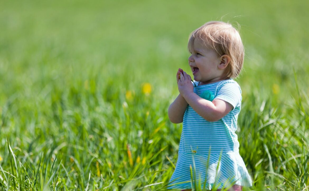 Young child in a blue striped outfit bringing their hands together in a grassy field.