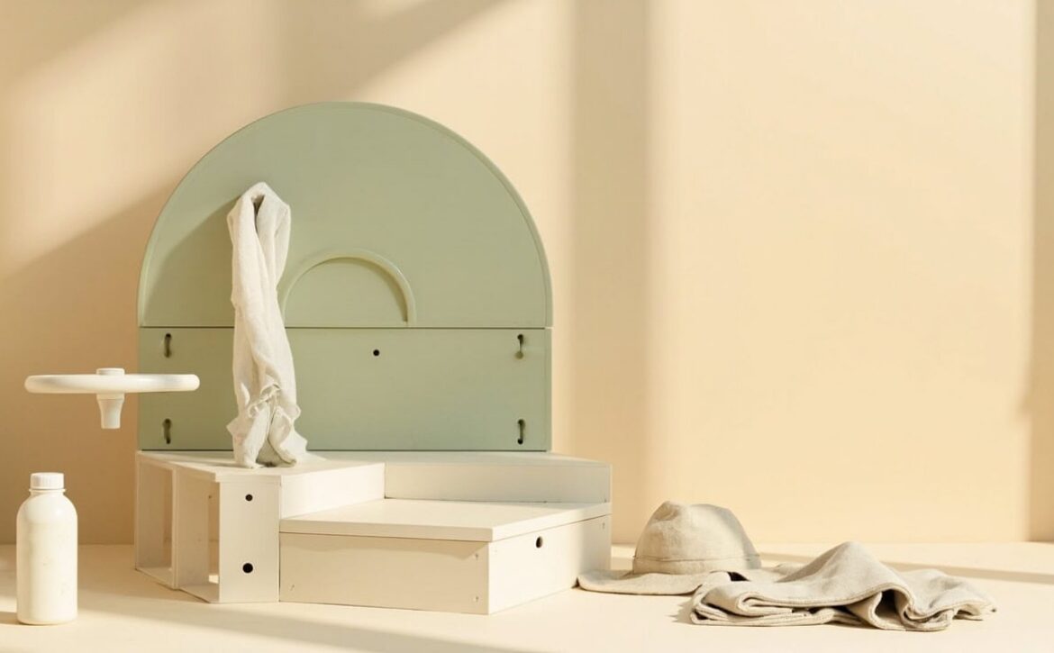 A warm cream still life shows a sage arched structure, white stepped blocks, a bottle, a sun hat and folded fabric.