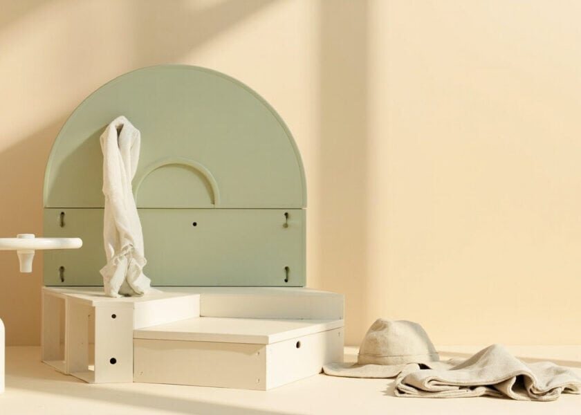 A warm cream still life shows a sage arched structure, white stepped blocks, a bottle, a sun hat and folded fabric.