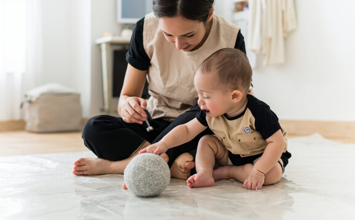 A caregiver leans toward a seated baby who reaches to a soft grey ball on a clear mat in a bright room.