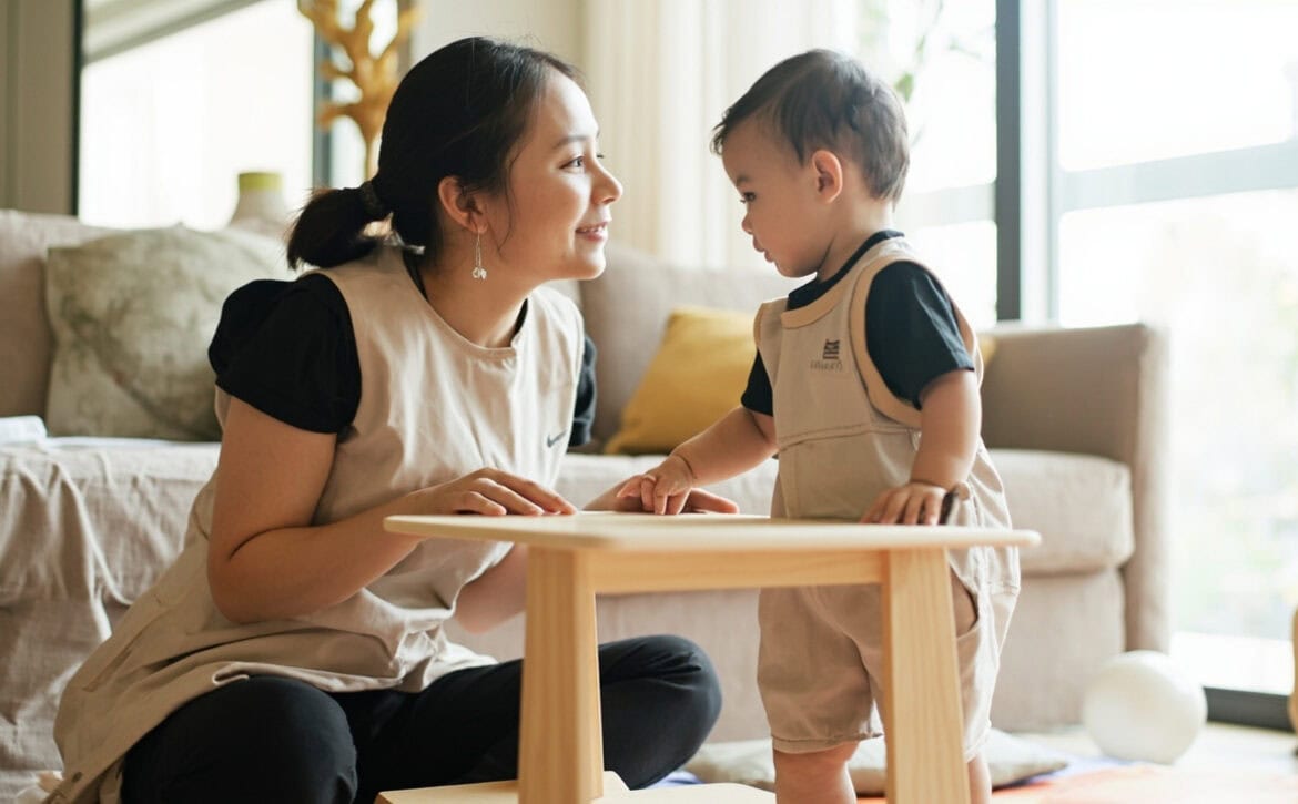 A caregiver kneels beside a baby standing at a low wooden table with both hands on top, in a bright living room with a smooth ball on a mat.