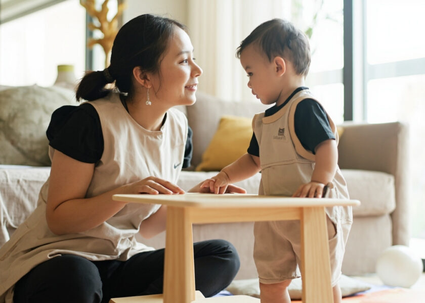 A caregiver kneels beside a baby standing at a low wooden table with both hands on top, in a bright living room with a smooth ball on a mat.