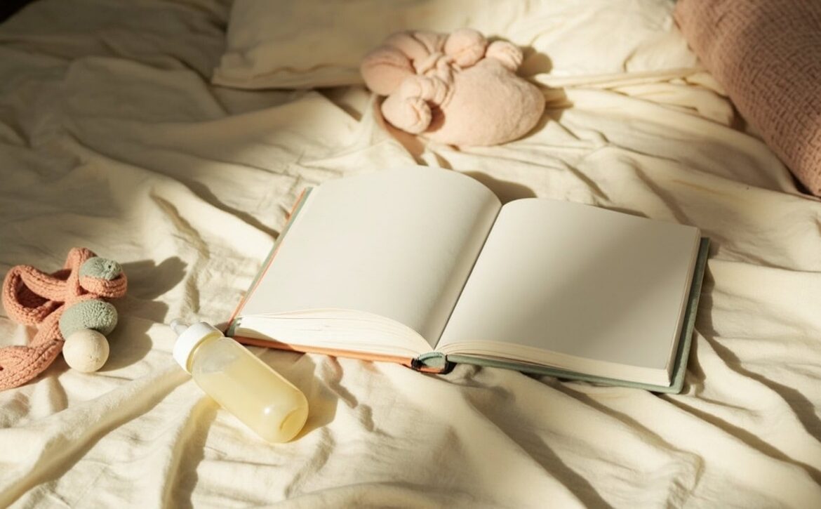 An open blank book, unbranded baby bottle and soft toys rest on rumpled cream bedding in warm evening light.