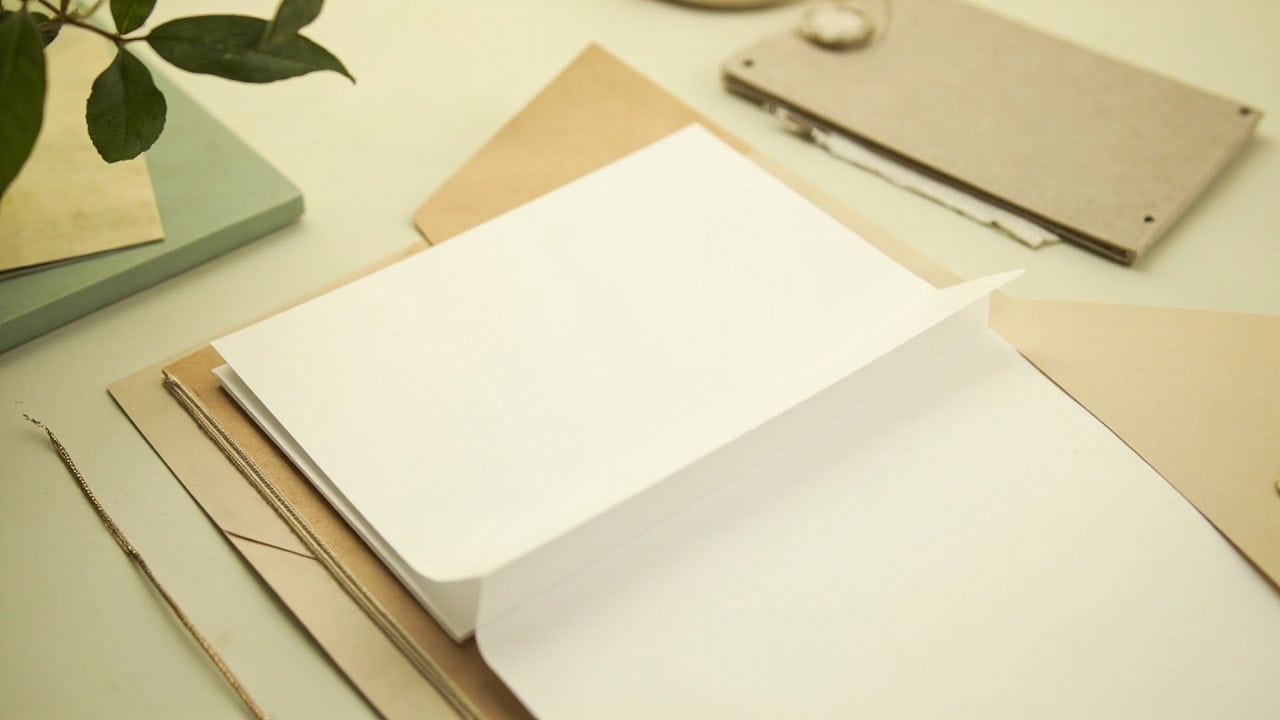 An open kraft folder holds blank cream papers on a warm tabletop, with muted green accents.