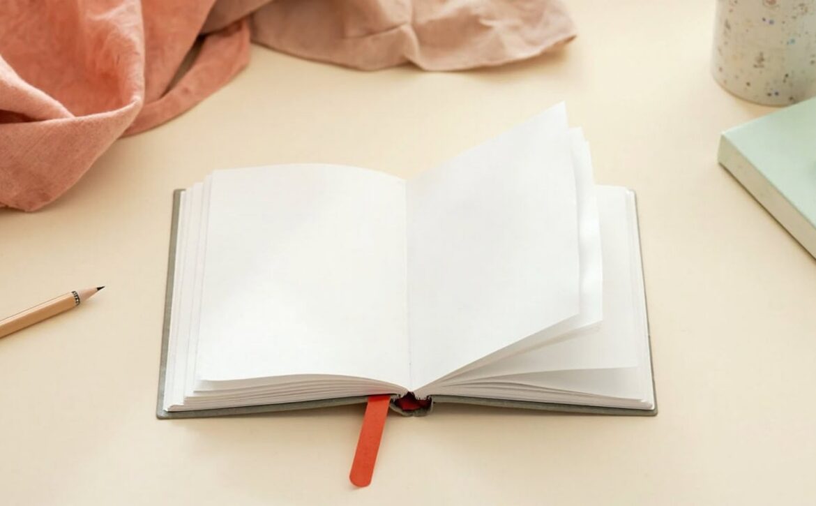 An open blank booklet rests on a cream table beside a wooden pencil, coral fabric, a speckled cup and a closed sage book.