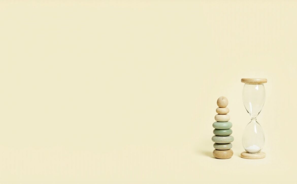Minimal ivory still life of a muted wooden stacking toy beside a clear hourglass, with broad empty space.