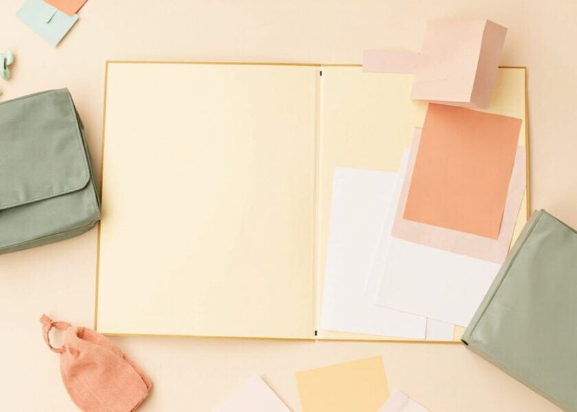 Overhead still life of an open blank cream folder, loose papers, muted sage bags and a small coral pouch.