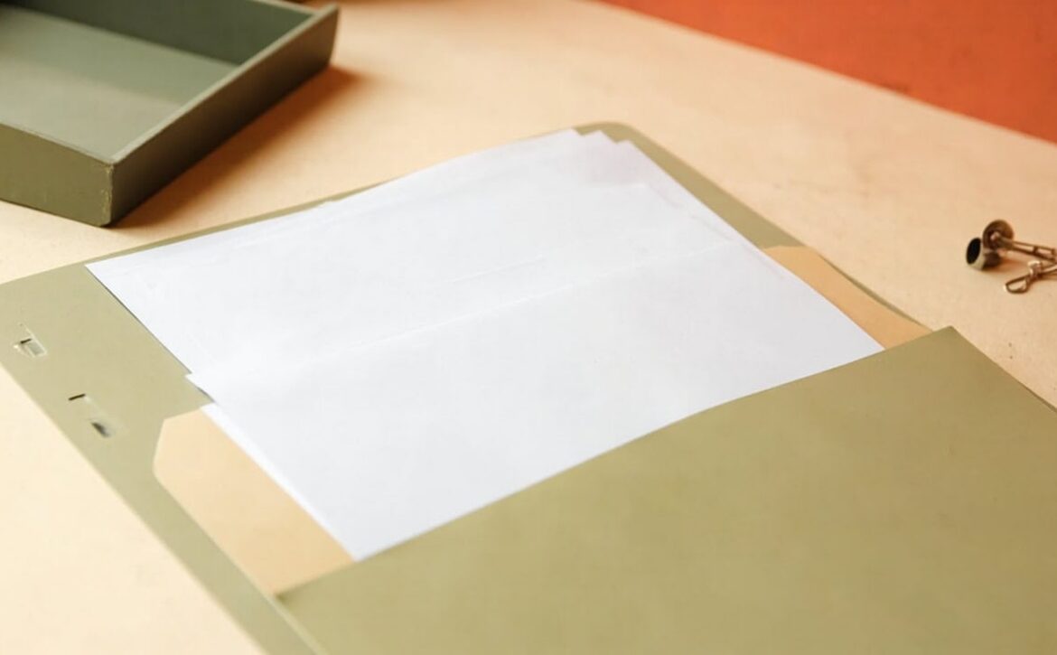 Open sage folder with blank white papers on a warm cream desk, beside a muted tray and several metal paperclips.