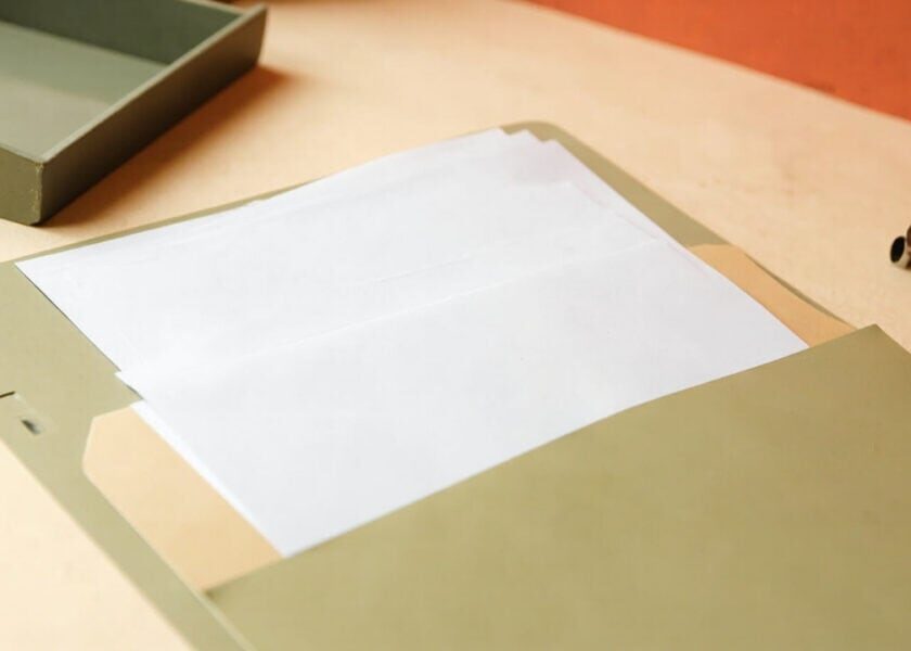 Open sage folder with blank white papers on a warm cream desk, beside a muted tray and several metal paperclips.