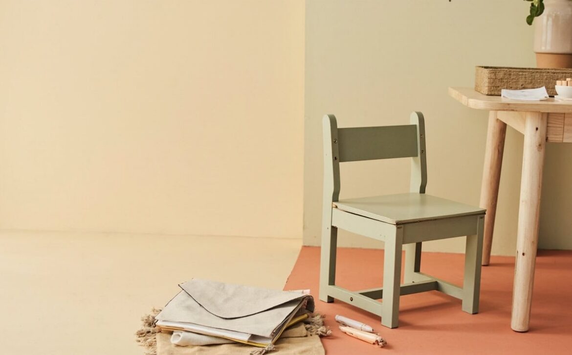 A small sage child-sized chair beside a wooden table, blank papers and craft materials in a warm, uncluttered room.