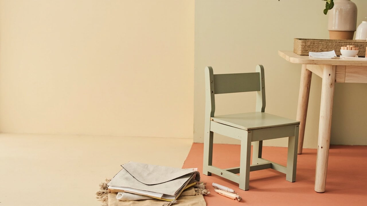 A small sage child-sized chair beside a wooden table, blank papers and craft materials in a warm, uncluttered room.