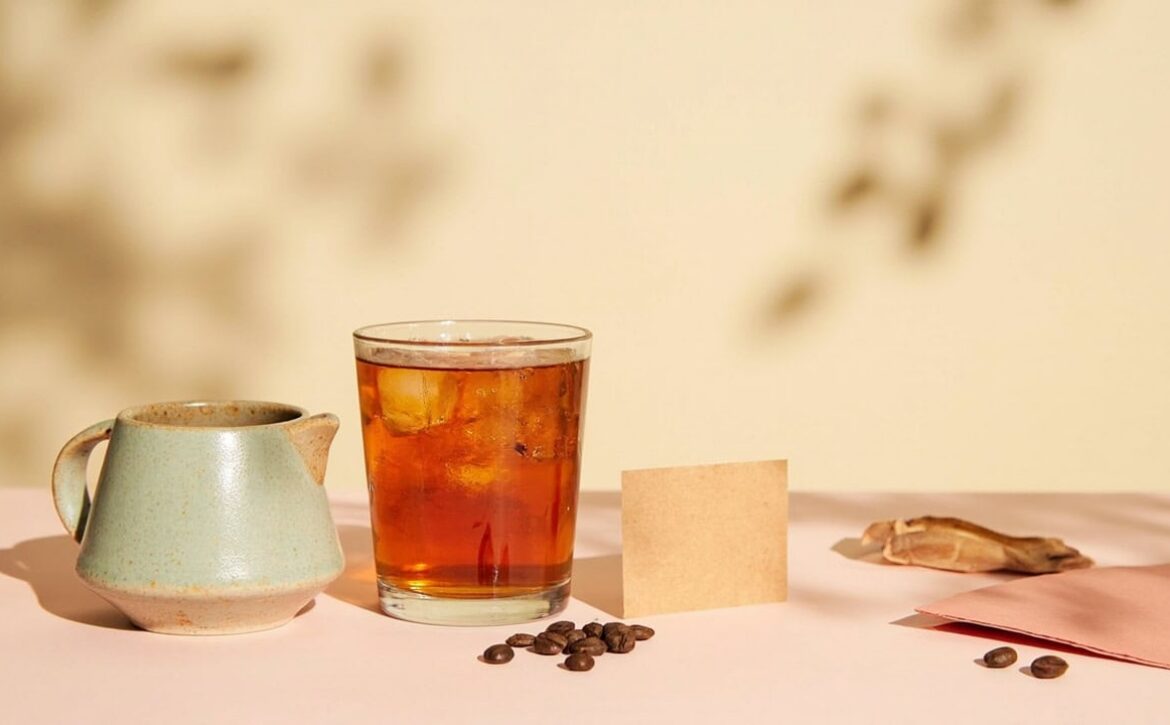 Warm still life of amber iced tea, a sage ceramic jug, coffee beans, loose leaves and blank paper cards.
