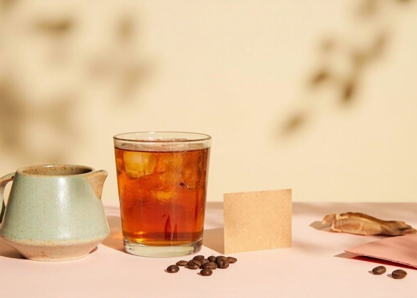 Warm still life of amber iced tea, a sage ceramic jug, coffee beans, loose leaves and blank paper cards.