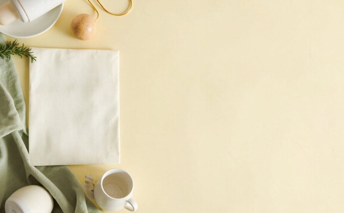 Overhead cream still life with a blank folded sheet, baby bottle, ceramic cup, sage fabric and a small wooden rattle.