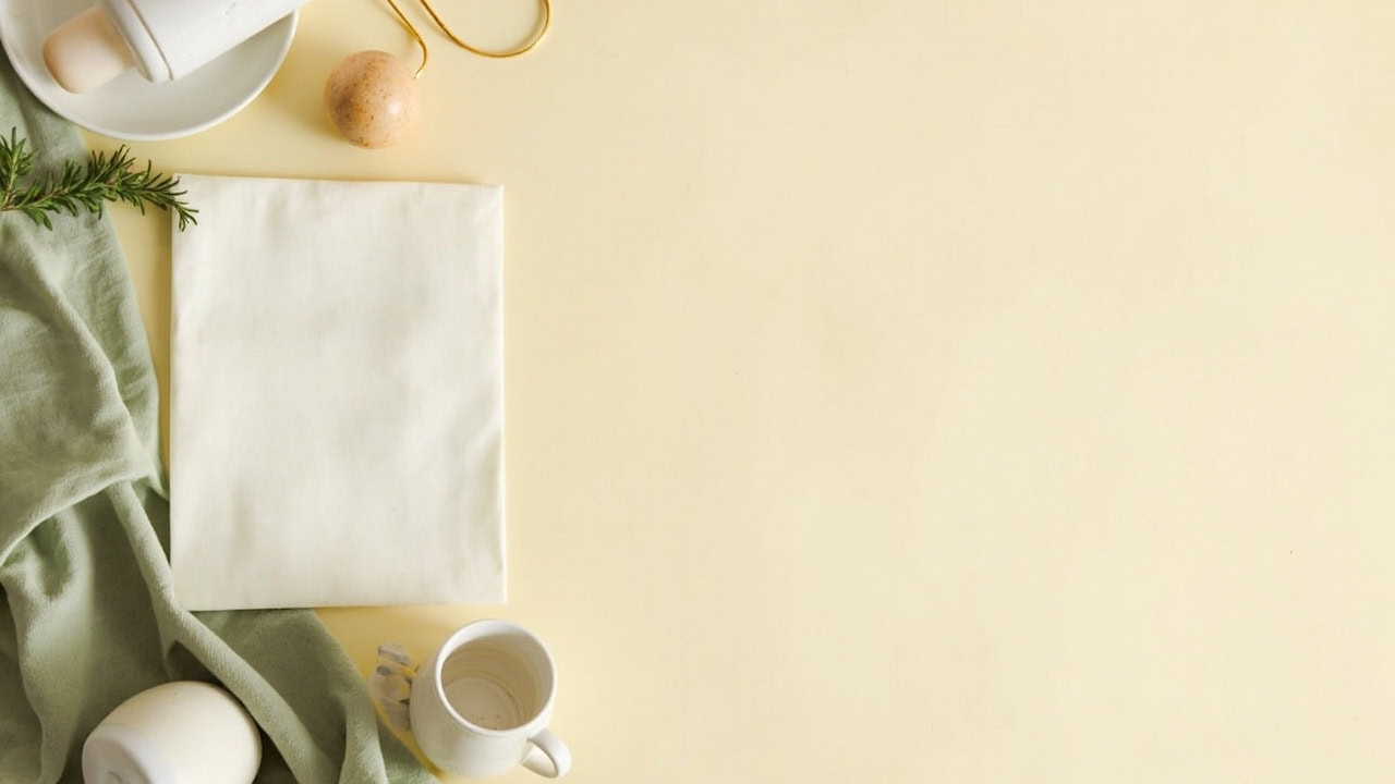 Overhead cream still life with a blank folded sheet, baby bottle, ceramic cup, sage fabric and a small wooden rattle.