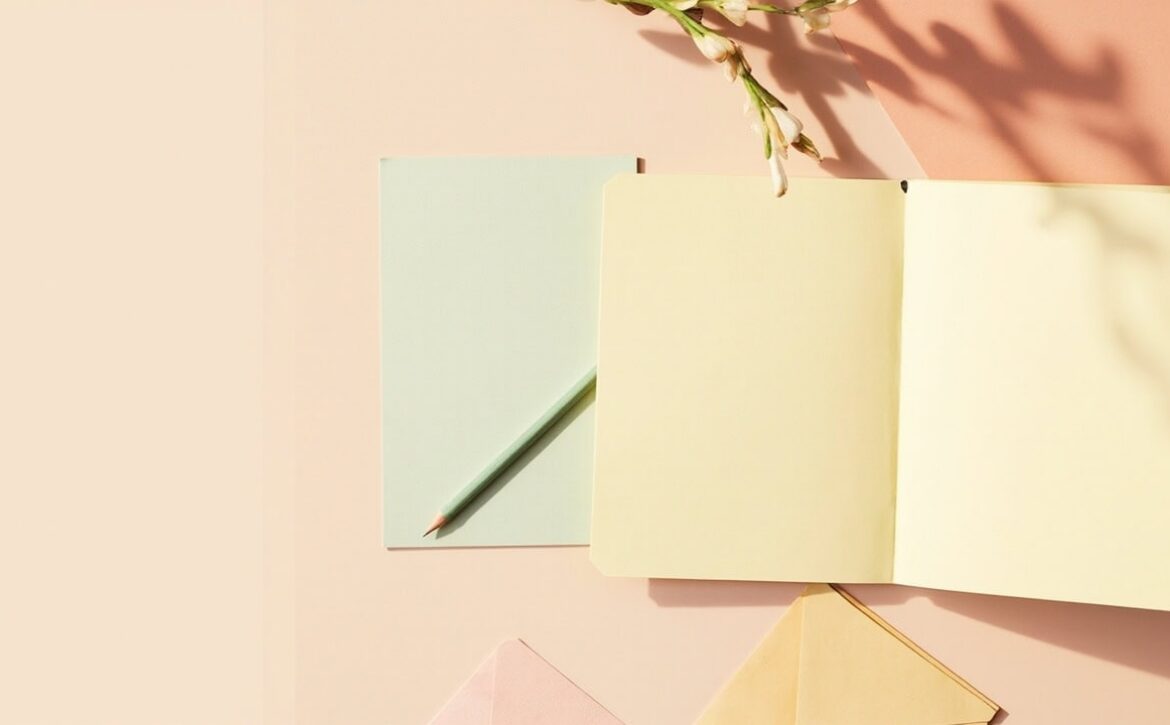 Overhead still life of a blank cream folder, sage card and pencil, with flowers and a partly cropped envelope in warm light.