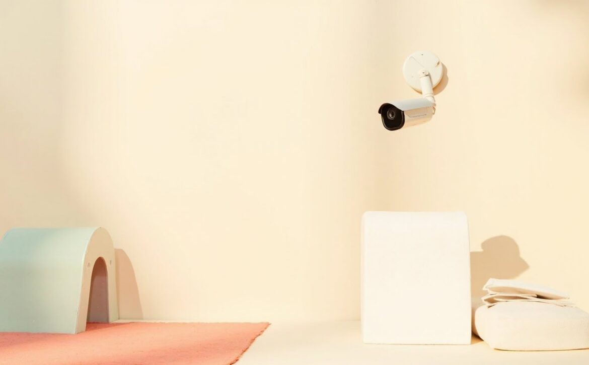 A wall-mounted security camera overlooks a sparse cream play area with a mint arch, white block and folded linens.