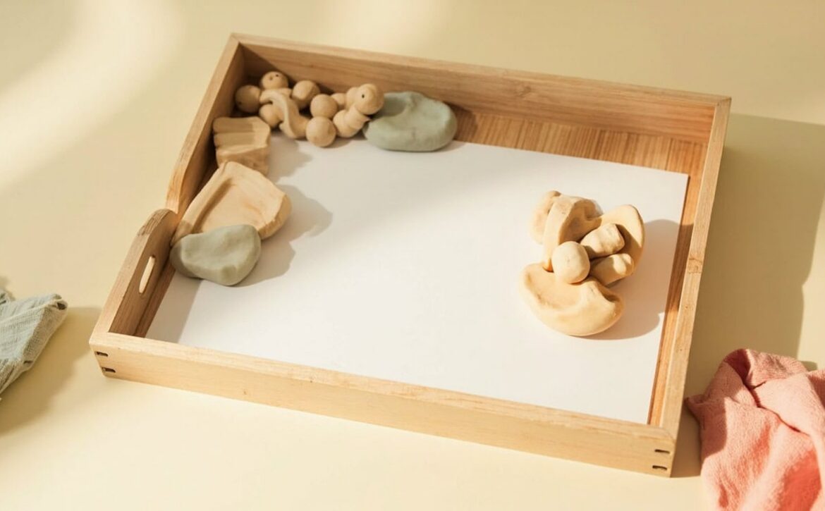 A wooden tray with blank paper, small wooden shapes and soft clay on a cream table, bordered by folded fabric.