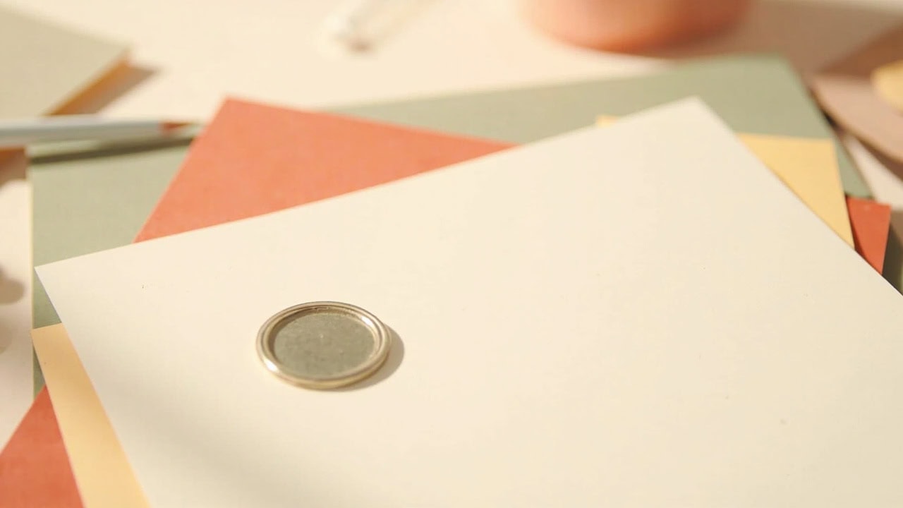 A plain silver badge rests on a cream document, with muted coral, yellow and sage papers arranged beneath in soft light.