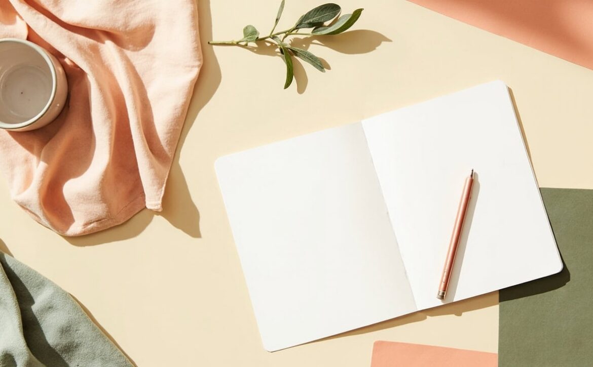 Overhead still life of a blank notebook and pencil on a cream tabletop, with a cup, folded fabrics and a green sprig.