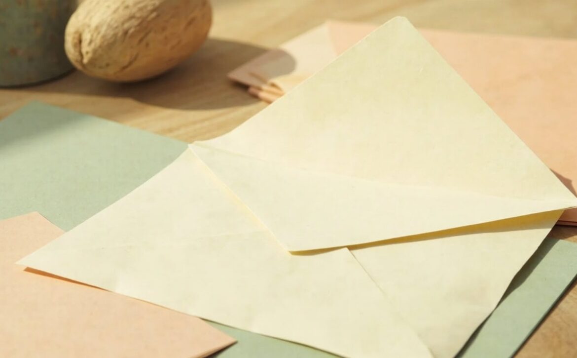 A blank cream envelope rests among muted folders and cards on a pale wooden table in warm daylight.