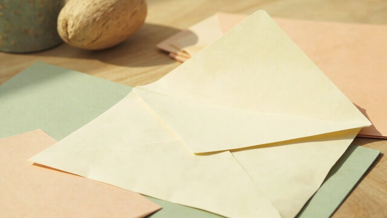 A blank cream envelope rests among muted folders and cards on a pale wooden table in warm daylight.