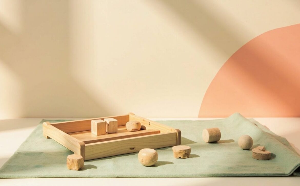 A wooden tray of unmarked blocks and round play pieces on a folded sage-green mat, framed by warm cream and coral.
