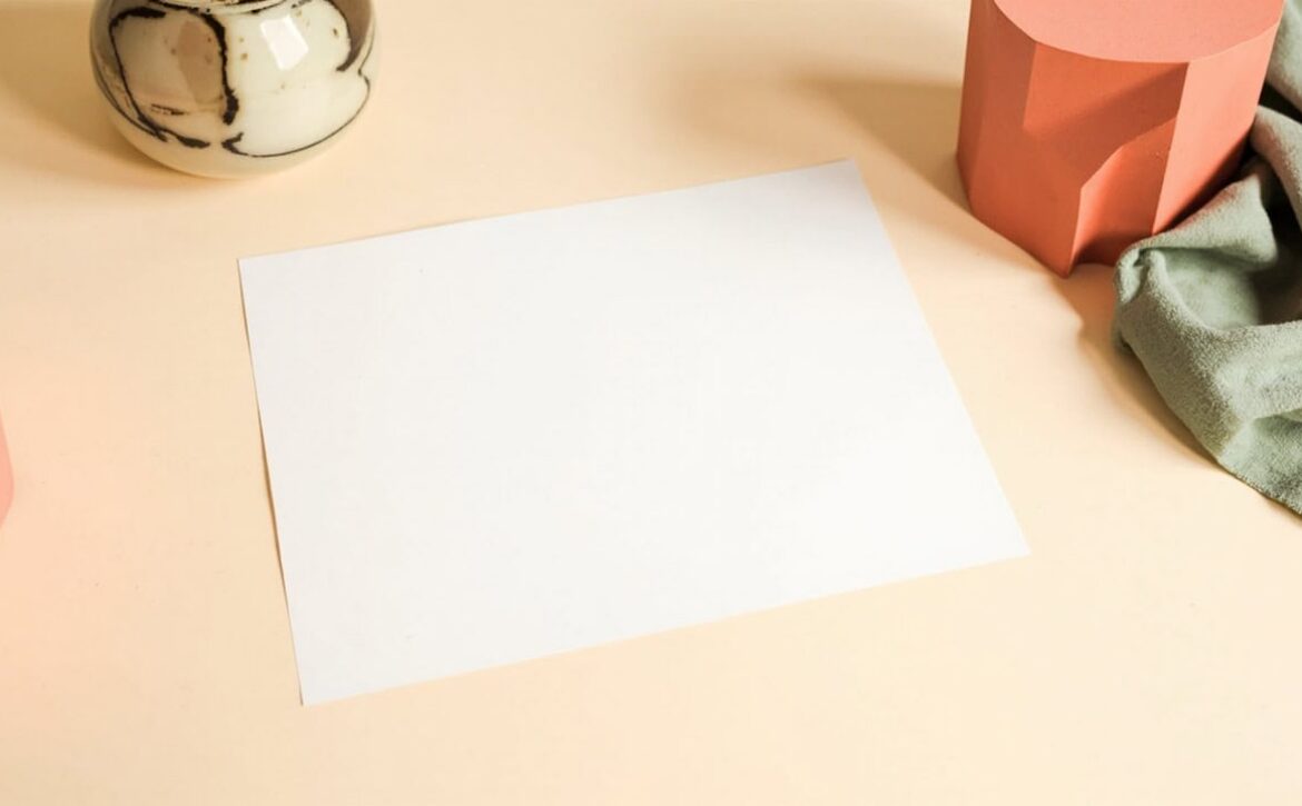 Blank paper on a cream desk, framed by a ceramic vessel, sage fabric and coral geometric forms.