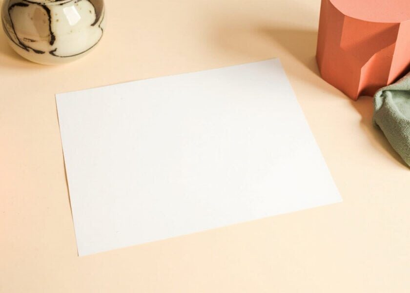 Blank paper on a cream desk, framed by a ceramic vessel, sage fabric and coral geometric forms.