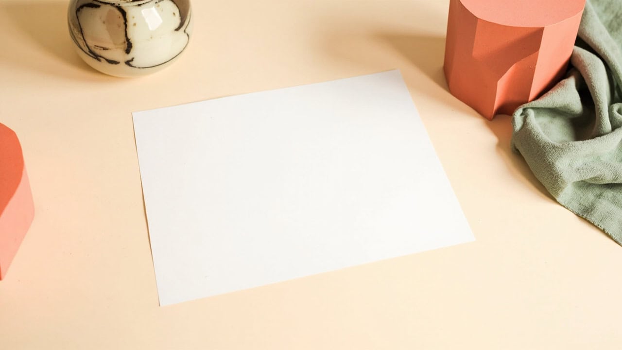 Blank paper on a cream desk, framed by a ceramic vessel, sage fabric and coral geometric forms.