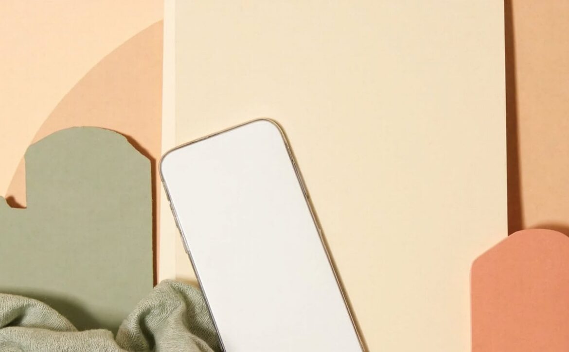 A blank smartphone rests on sage fabric beside a cream folder and muted abstract arch-shaped forms.