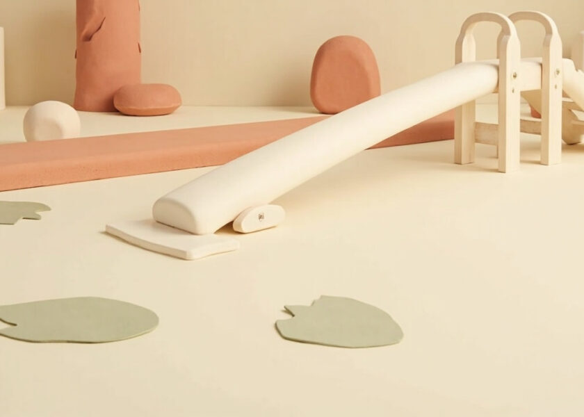 A cream-and-coral still life of an unmarked low balance beam, small climbing frame and slide, with leaf-shaped stepping pads.