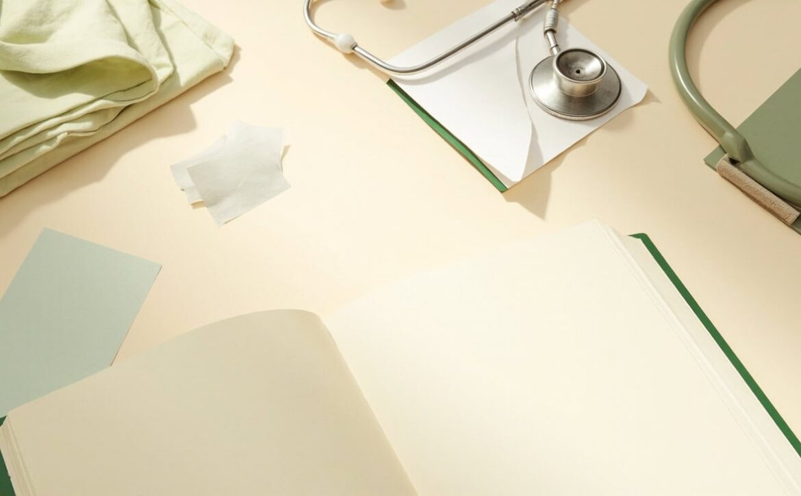 An open blank notebook, stethoscope, folded cloth and muted green stationery on a cream tabletop.