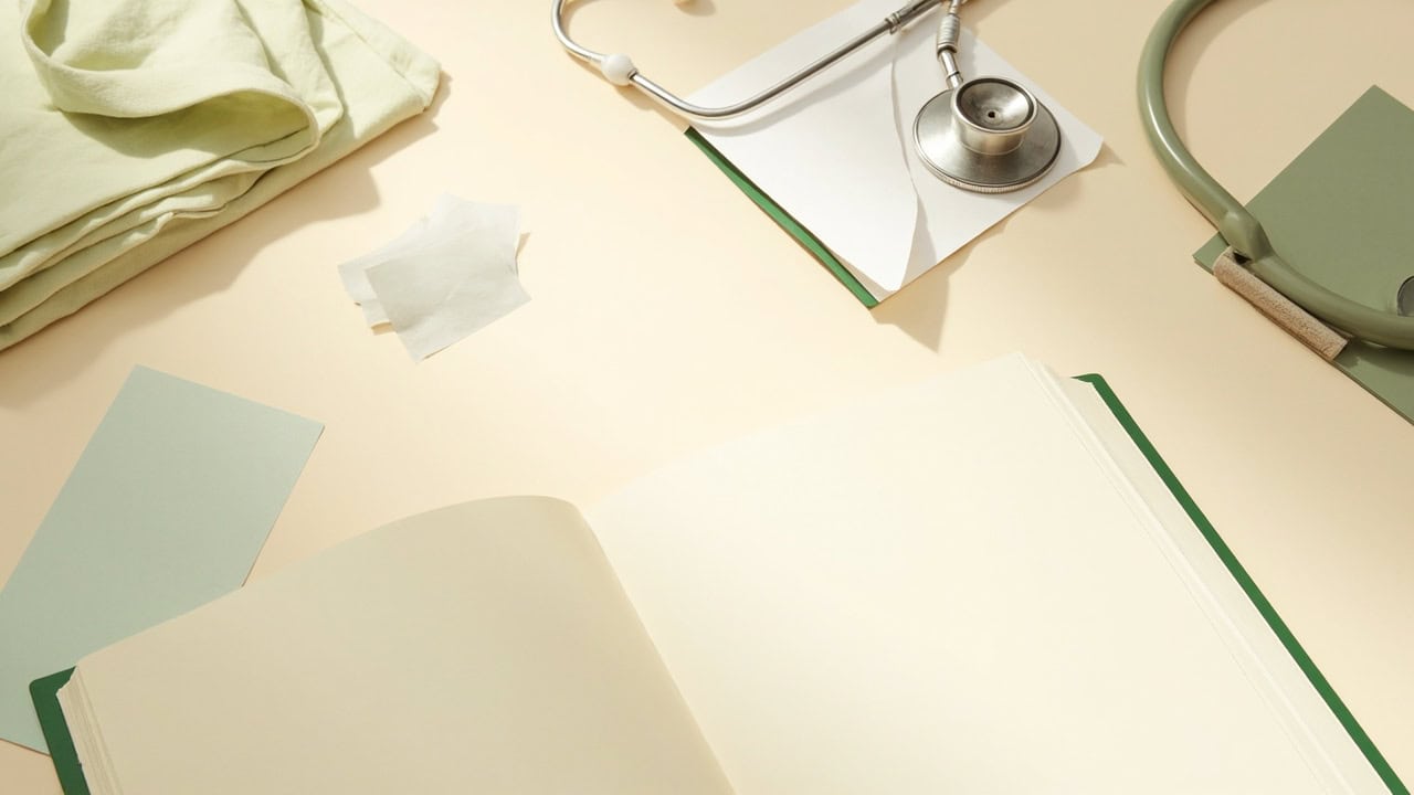 An open blank notebook, stethoscope, folded cloth and muted green stationery on a cream tabletop.