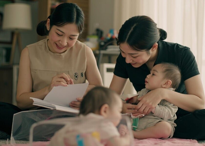 Disclosed AI illustration of a parent and childminder reviewing a plain note while an awake infant plays safely in a Singapore living room