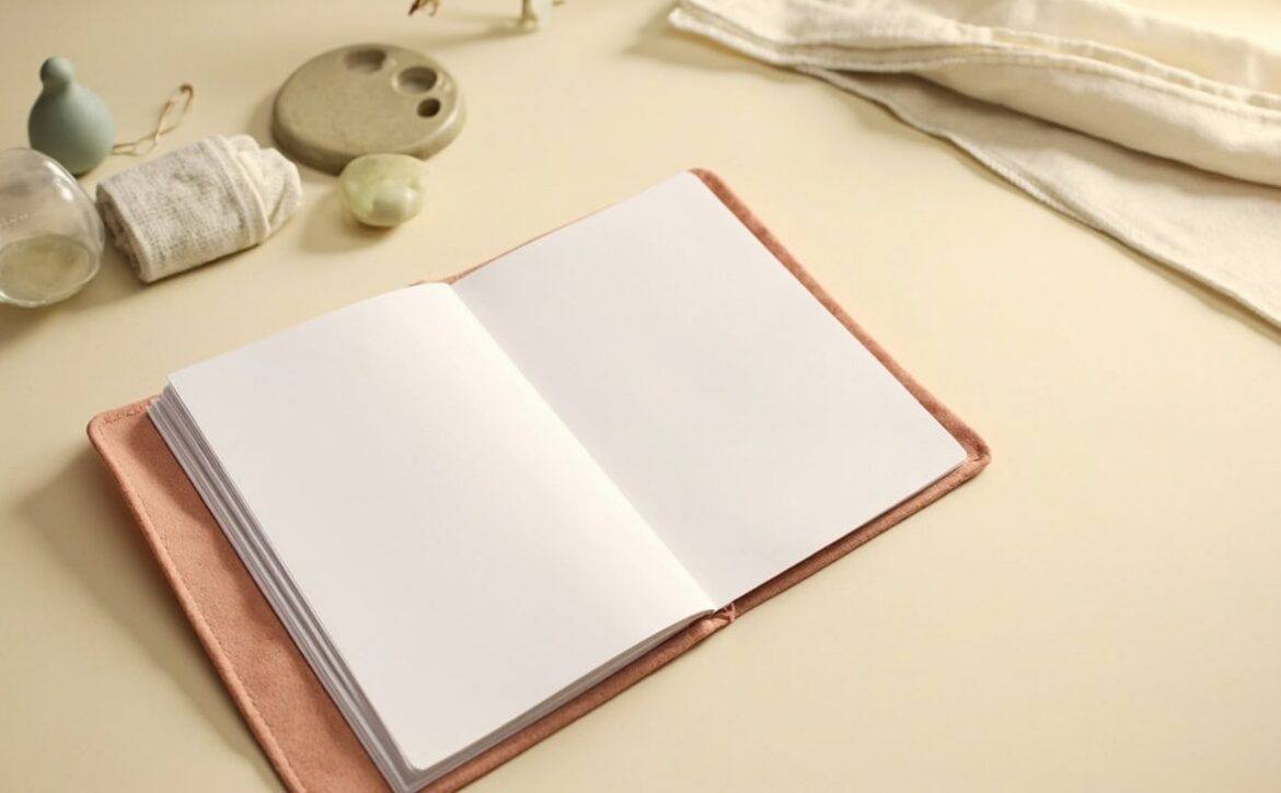An open blank notebook on a coral folder, surrounded by folded cloths and small glass, ceramic and sage objects.