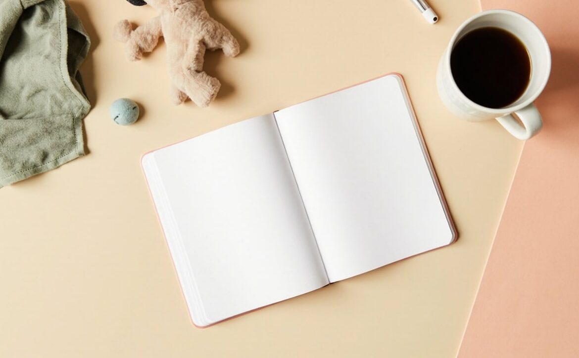 Overhead still life of a blank notebook, soft toy, folded cloth and cup on a warm cream-and-coral desk.