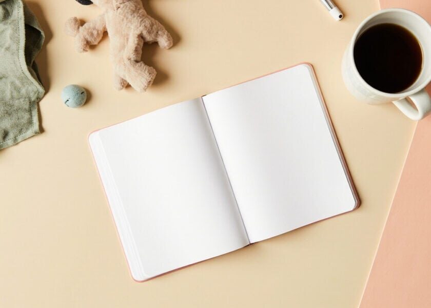Overhead still life of a blank notebook, soft toy, folded cloth and cup on a warm cream-and-coral desk.