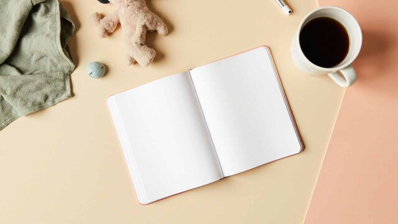 Overhead still life of a blank notebook, soft toy, folded cloth and cup on a warm cream-and-coral desk.