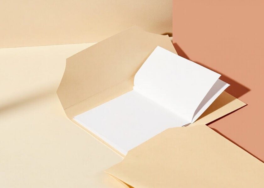 Open plain folder with blank white sheets and neutral envelopes on a warm cream and coral tabletop.