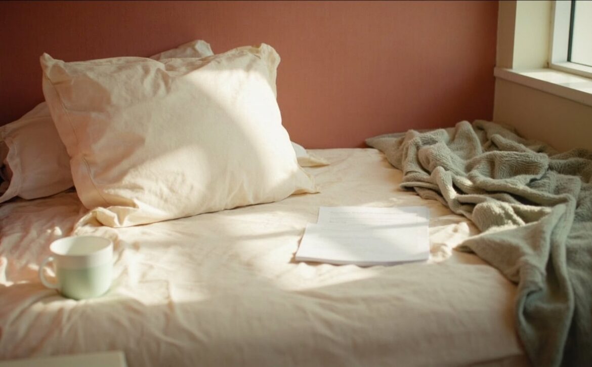 A sunlit bed with cream pillows, a rumpled sage throw, a plain mug and unmarked papers.