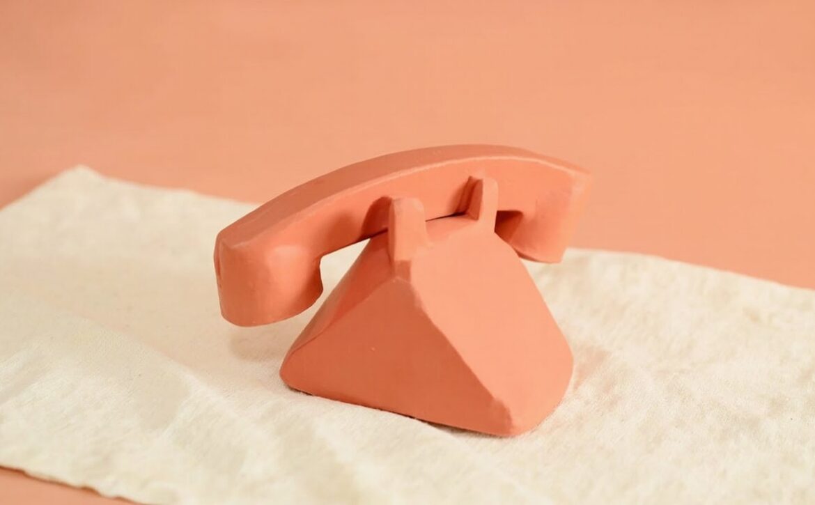 A coral clay, old-fashioned telephone form with a curved receiver rests on cream linen against a peach background.