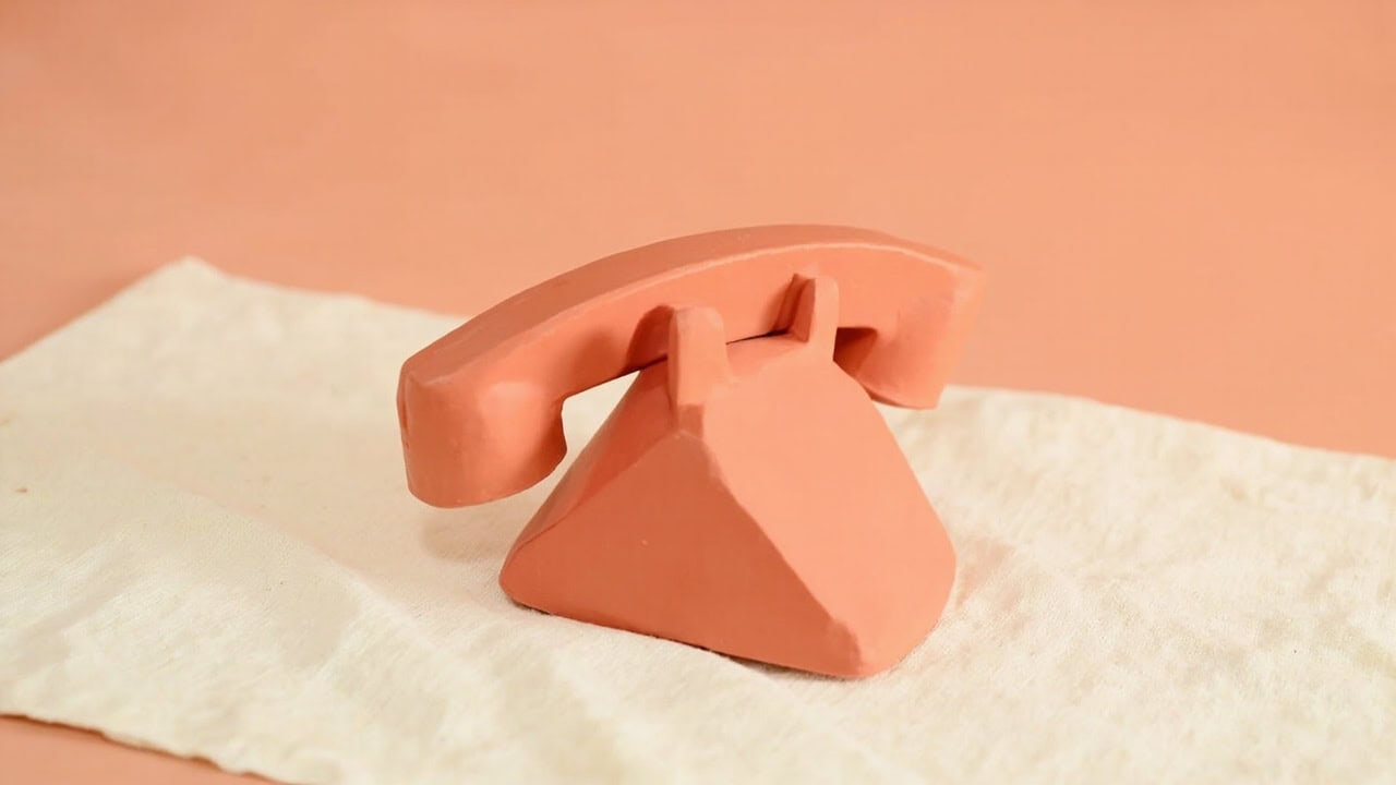 A coral clay, old-fashioned telephone form with a curved receiver rests on cream linen against a peach background.