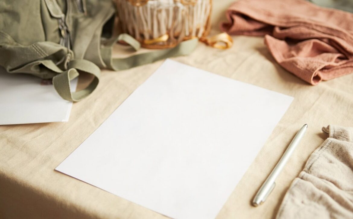A blank white sheet rests on a cream table beside a silver pen, a small backpack, a basket and folded clothes.